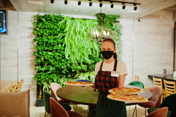 Waiter in protective mask hold pizza in pizzeria.