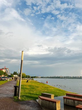 Tarnopil, Ukrayna - 10 Ağustos 2025: Yeşil çimenler, banklar ve uzak şehir manzaralı huzurlu göl kenarı geçidi.