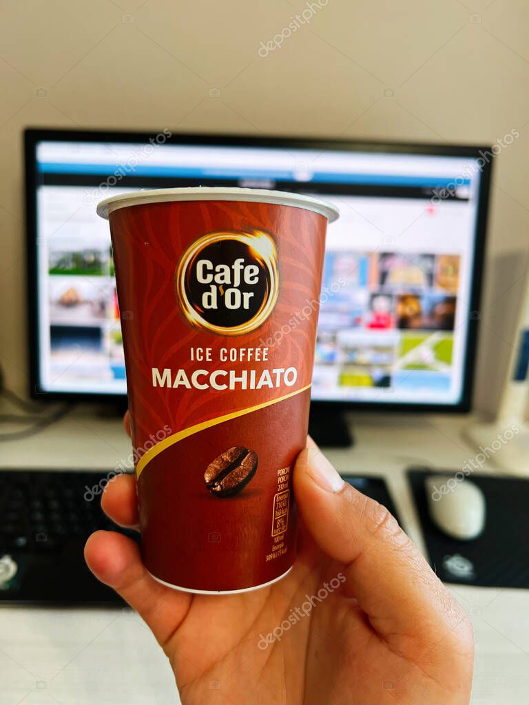 Kyiv, Ukraine - August 10, 2025: A person holding Cafe d'Or macchiato iced coffee cup near a workstation with a computer.