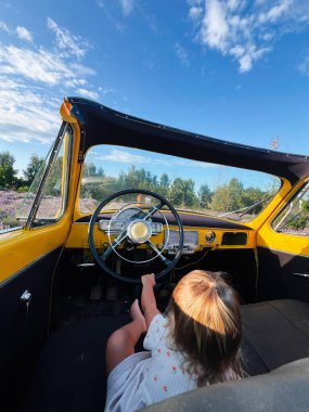 Bir çocuk, mavi gökyüzüne karşı klasik sarı bir arabanın içinde oturuyor..