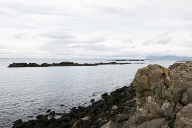 İzlanda 'da kayalık kıyı şeridi. Yumuşak bulutların altında uzak, sakin adalar görünüyor.