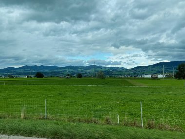 Etrafı dağlarla çevrili yeşil otlaklar bulutlu bir gökyüzünün altında, İsviçre kırsalını gösteriyor..