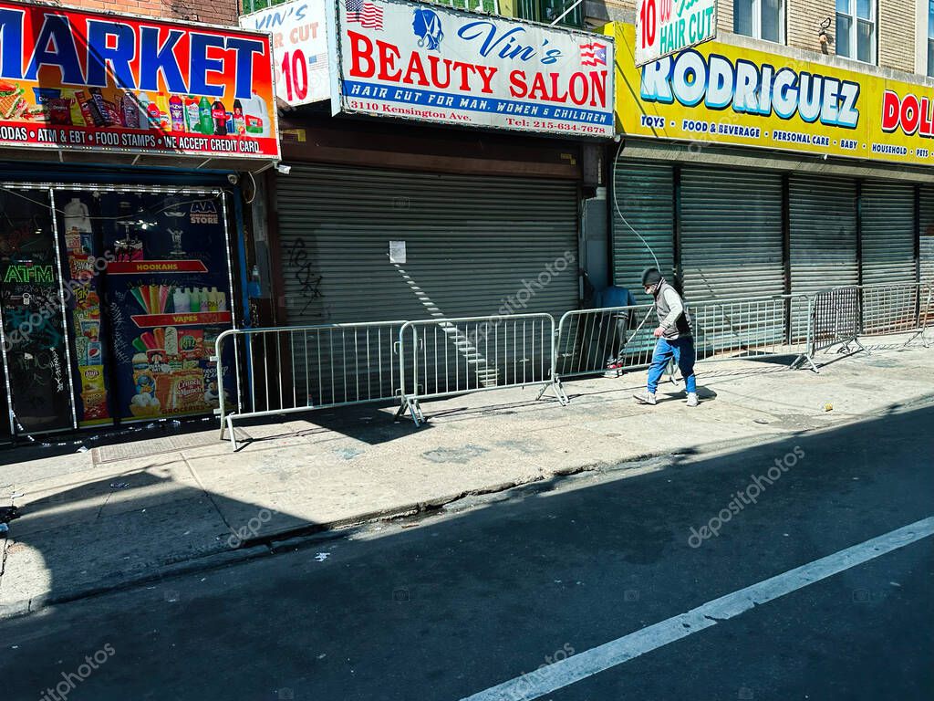 Philadelphia, Pennsylvania, USA - April 29, 2025: A Zombieland, Kensington Ave, Philadelphia street scene showing closed storefronts, bold market signs, and a man walking past barriers.