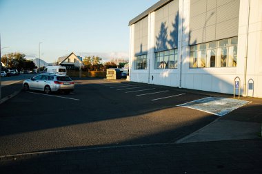Akranes, İzlanda - 17 Eylül 2025: Akranes, İzlanda 'da güneşli park alanı, büyük beyaz sanayi binası, park edilmiş arabalar ve uzun gölgeler.