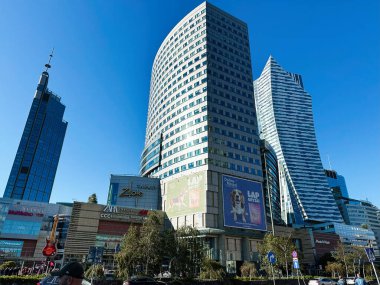 Varşova, Polonya - 19 Ekim 2024: Yüksek cam binalar, işlek caddeler ve Polonya 'nın Varşova şehrinde açık mavi gökyüzü altında alışveriş merkezi tabelaları bulunan canlı şehir manzarası.