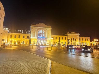 Tarnow, Polonya - 25 Ekim 2024: Sıcak sokak ışıkları, kaldırım taşı meydanı ve hareket halindeki araçlarla dolu Tarnow istasyonunda canlı bir gece sahnesi.