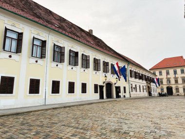 Zagreb, Hırvatistan - 23 Ekim 2024: Pencereleri kapatılmış soluk sarı bir cephe Zagreb, Hırvatistan ve AB bayrakları arasında dalgalanan kaldırımlı bir sokak, Hırvat ve AB bayrakları hizasında.