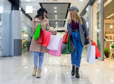 İki genç arkadaş, sarışın ve esmer alışveriş merkezinde eğleniyorlar, yürüyorlar, sohbet ediyorlar, gülüyorlar ve alışveriş çantalarını taşıyorlar.