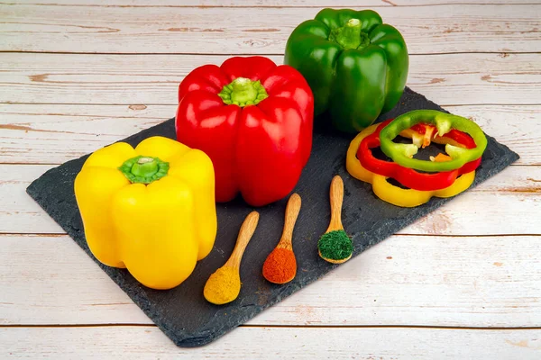 Tam ve dilimlenmiş kırmızı, yeşil, sarı biber ve siyah taş masada baharatlı üç tahta kaşık.