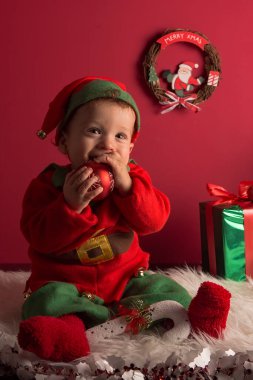 Tatlı bir çocuk Elf gibi giyinmiş beyaz halıda oturuyor ve Noel süsü takmış hediyelerle.