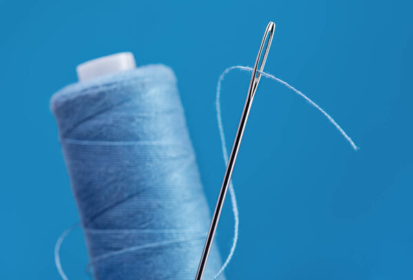 Большая серебряная игла для шитья ниткой. Вертикально стоящая катушка синих нитей. Синий фон, пространство для копирования
.