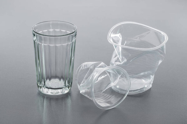 Simple glass and crumpled plastic cup on gray background, close-up. Recycling concept.