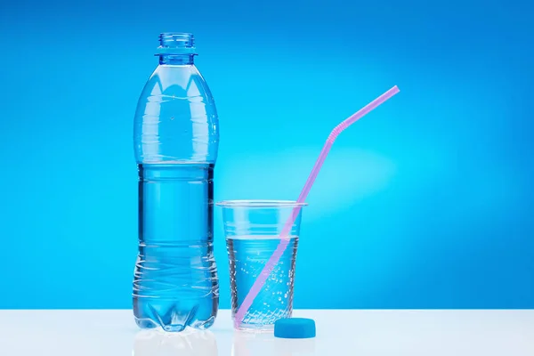 Water with gas in plastic glass with pink straw. Bottle with fresh water on white table and blue background. Copy space.