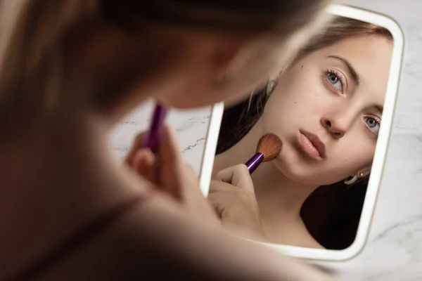 Blonde girl at  mirror, makeup brush in hand. To do makeup. Reflection in mirror.