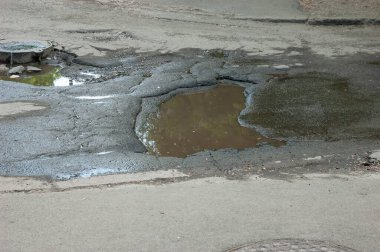 Su birikintileri ve çatlakları olan, arka planda rögar deliği olan hasarlı bir yol..