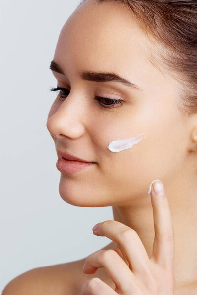 Skin care. Lady putting face cream. Photoset of attractive brunette girl on grey background. Cute girl preparing to start her day. She is applying moisturizer cream on face.