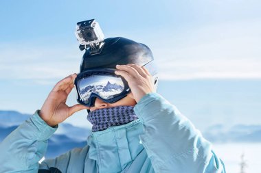 Kayakçı ile eylem fotoğraf makinesi üzerinde bir kask. Kayak kar yağdı dağlara yansıması ile gözlükler. Adamın dağlar ve mavi gökyüzü arka plan üzerinde Kayak Merkezi, portre. Kayak gözlüklü