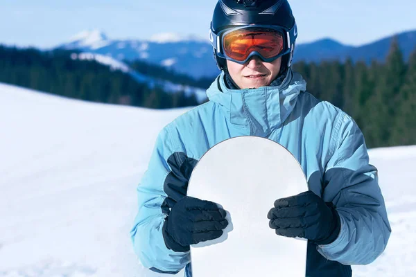 Snowboarder with action camera on a helmet. Ski goggles  with the reflection of snowed mountains. Portrait of man at  ski resort on the background blue sky,  hold snowboard. Wearing ski glasses