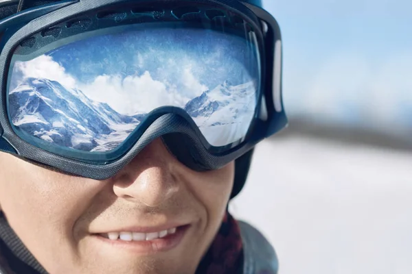 Close up of the ski goggles of a man with the reflection of snowed mountains.  A mountain range reflected in the ski mask.  Man  on the background blue sky. Wearing ski glasses. Winter Sports.