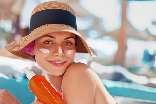 Young woman with sun cream on face holding sunscren bottle on the beach. Female in hat applying  moisturizing lotion on skin.