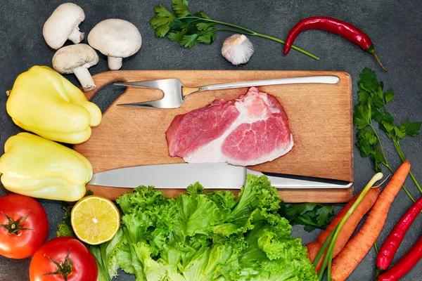 Koyu arka planda çiğ et sağlıklı yemek için taze malzemelerle çiğ organik sebzeler. Biberiye ve baharatlar ile kesme tahtası üzerinde çiğ sığır eti biftek. Üstte görüntü.