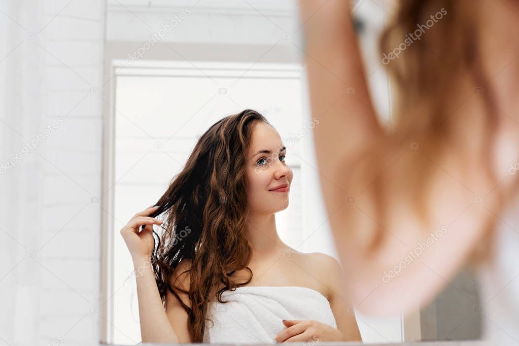 Hair and body care. Woman touching wet hair and smiling while looking in the mirror. Portrait of girl  in bathroom applying conditioner and oil.Portrait of female uses protection moisturizing cream.