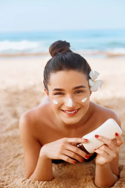 Beautiful Woman smile applying sun cream on face. Skin and  Body  care. Sun protection. Girl in bikini holding sunscreen bottle on the beach. Female in hat smear moisturizing lotion on skin