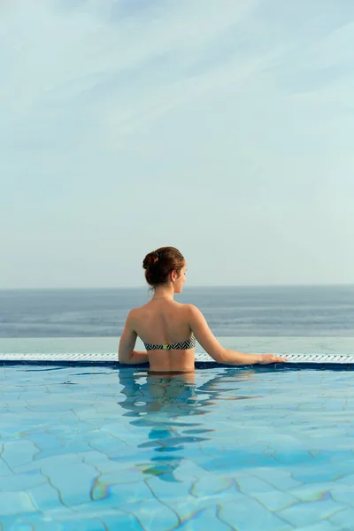 Luxury Resort. Woman Relaxing In Infinity Swimming Pool Water. Beautiful Happy Healthy Female Model Enjoying Summer Travel Vacation, Looking At Sea View. Summertime Recreation, Relax And Spa Concept
