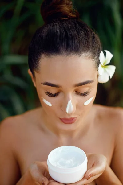 Beauty face. Woman model with natural makeup and healthy skin. Portrait of beautiful girl with smile holding cosmetic cream in tropical nature