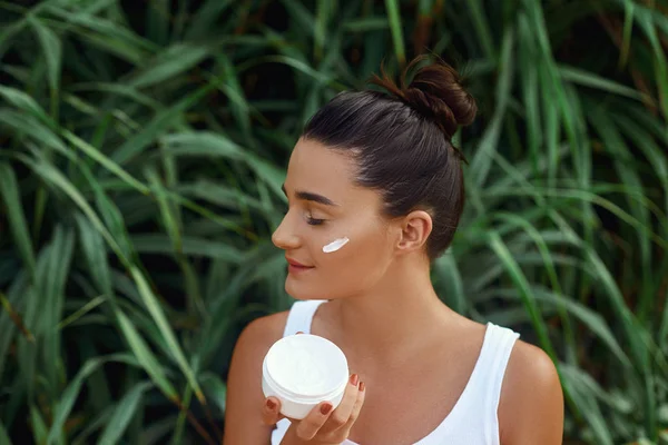 Beauty Woman Concept. Skin care.  Portrait of female model  holding and applying cosmetic moisturizing cream and touch own face. Nude make-up. Skin Protection  in tropical nature