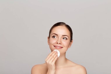 Healthy fresh girl removing makeup from her face with cotton pad. Beauty woman cleaning her face with cotton swab pad isolated on grey background. Skin care and beauty concept. clipart