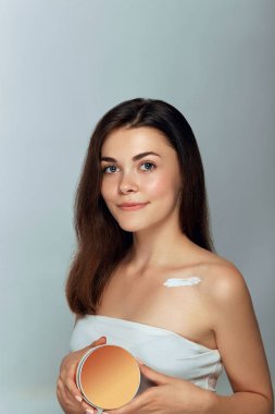 Cilt bakımı. Güzellik Konsepti. Kozmetik krem tutan genç ve güzel bir kadın. Yumuşak cilt ve çıplak omuzlar, açık renk makyajlı bir model, elinde nemlendirici krem tutan ve kendi yüzüne dokunan bir kadın portresi. Cilt Koruma ve Dermatoloji.