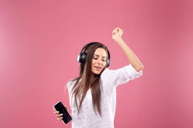 Kablosuz kulaklıklar takan, müzik dinleyen, cep telefonu kullanan ve pembe arka planda dans eden güzel genç bir kadın. Kız kablosuz kulaklık kullanıyor.