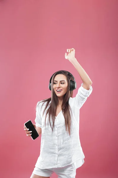 Beautiful young woman in wireless headphones listening to music using mobile phone and dancing on pink background. Girl uses wireless earphones