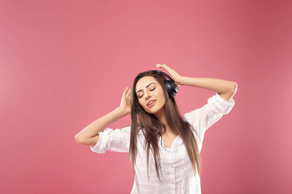 Young woman on pink background listening to music with wireless headphones. Girl using phone of listening to music with wireless earphones.
