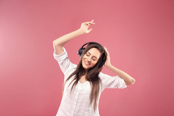 Beautiful young woman in wireless headphones listening to music and dancing on pink background. Girl uses wireless earphones