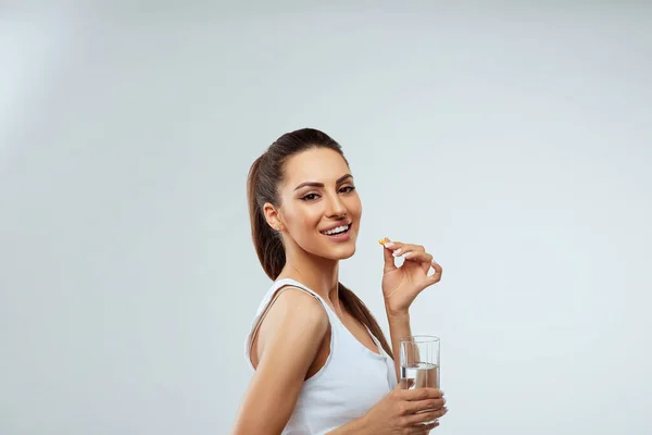 Gülümseyen kadın sabahları vitamin hapları alıyor ve elinde bir bardak taze su tutuyor. D vitamini, E vitamini, balık yağı kapsülleri, Omega-3. Diyet olsun. Beslenmek. Sağlıklı Yeme