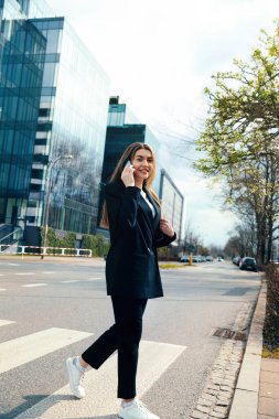 Confident young woman in a stylish outfit walks along a city street, talking on her smartphone with modern buildings in the background, showcasing urban lifestyle