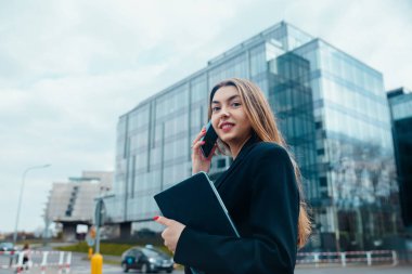 Confident young woman engaged in a phone conversation while holding a laptop in an urban environment with modern architecture reflecting her professional lifestyle