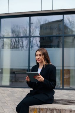 Young woman outdoors with a tablet, wearing a fashionable black coat, focused on her digital tasks, surrounded by contemporary architecture