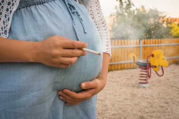 Pregnant woman smoking cigarette on the children playground - smoking addiction concept