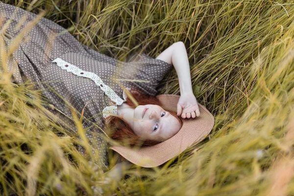 赤い髪の少女はエレガントなドレスで日没時に夏の牧草地に草の中に横たわっています — ストック写真