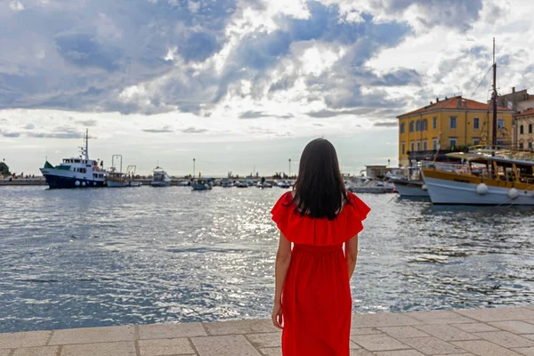 Kırmızı elbiseli kadın liman manzarasına bakıyor. Parlak kırmızı elbiseli bir kadın deniz kenarında geziniyor, dramatik bulutlu bir gökyüzünün altında limandaki teknelere bakıyor.