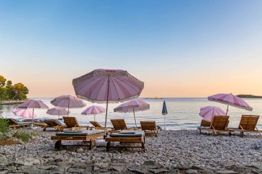 Gün batımında pembe şemsiyeli boş çakıl taşı plajı. Bir yaz akşamı gün batımında sakin denize bakan, tahta şezlonglar ve pembe şemsiyelerle dolu huzurlu çakıl taşı plajı.