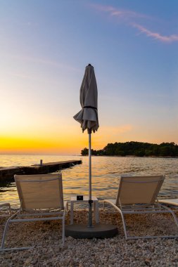 Gün batımına bakan boş plaj sandalyeleri. İki şezlong ve kayalık bir sahilde kapalı bir şemsiye, sakin deniz ve uzak ada üzerinde huzurlu bir günbatımına bakıyor.