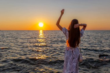 Genç kız okyanusun kenarında günbatımının tadını çıkarıyor. Parlak bir günbatımında deniz kenarında duran, barışçıl ve yansıtıcı atmosferi kucaklayan genç bir kızın arka görüntüsü.