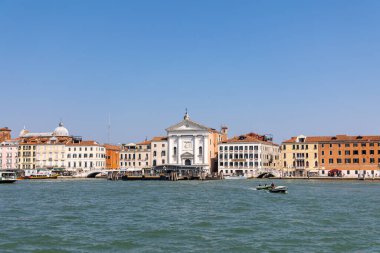 Venedik, İtalya, 11 Ağustos 2025: Venedik kıyısı, kilise ve renkli binalar. Beyaz bir kilise cephesi, renkli tarihi binalar ve kanalda gezinen teknelerin yer aldığı Venedik kıyısı manzarası..