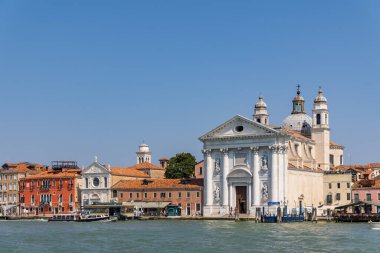 Venedik, İtalya, 11 Ağustos 2025 tarihi kilise ve kubbesi olan Venedik kıyısı. Venices rıhtımının klasik manzarası. Açık mavi gökyüzü altında kubbeleri ve renkli binaları olan büyük beyaz bir kilise..