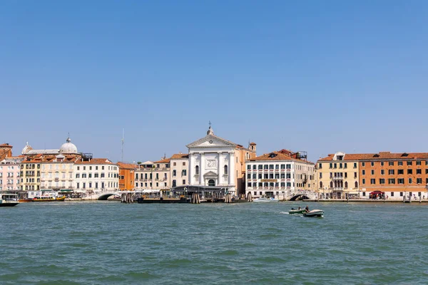 Venedik, İtalya, 11 Ağustos 2025: Venedik kıyısı, kilise ve renkli binalar. Beyaz bir kilise cephesi, renkli tarihi binalar ve kanalda gezinen teknelerin yer aldığı Venedik kıyısı manzarası..