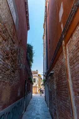 Venedik 'te güneşli bir sokağa açılan dar bir sokak. Tuğlalı sokak, turistlerin açık mavi gökyüzünün altındaki renkli binaların önünden geçtiği parlak bir Venedik caddesine çıkıyor.